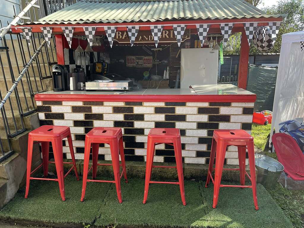 Outdoor backyard bar decorated with a racing checkerboard theme, featuring red bar stools and a brick-patterned counter.