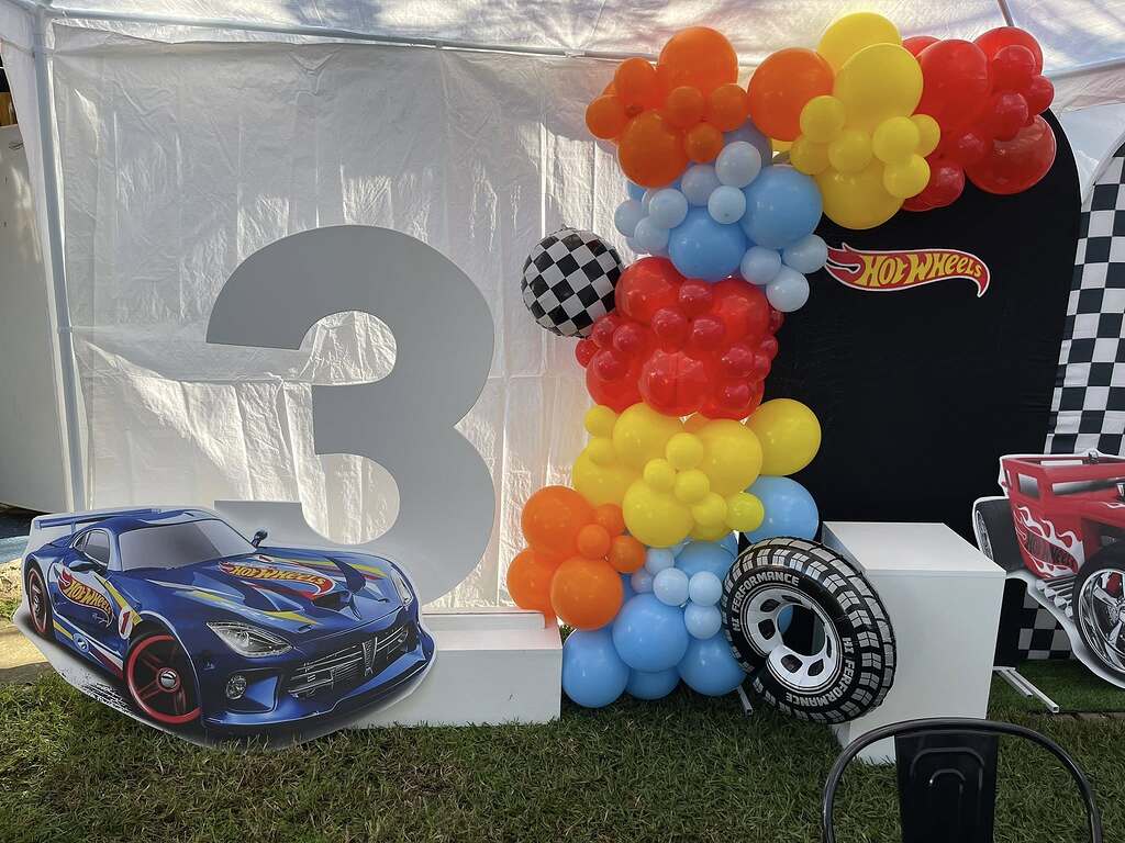 Hot Wheels themed third birthday party display with colorful balloon arch, number three prop, and racing car cutouts.