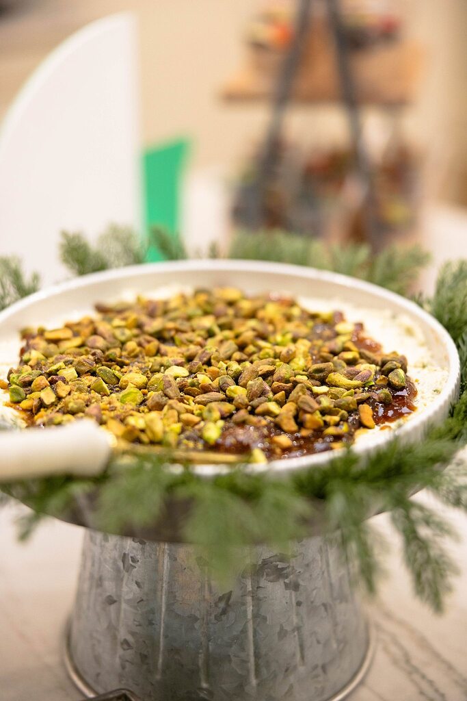 Creamy appetizer topped with jam and chopped pistachios on a pedestal stand with festive evergreen sprigs.