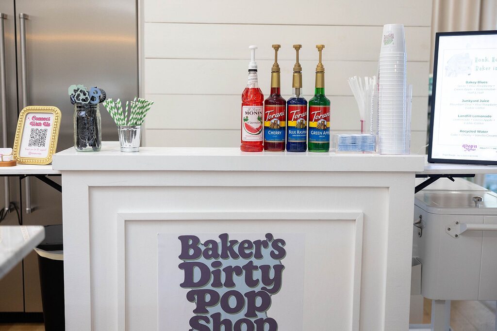 Tabletop drink station at Baker's Dirty Pop Shop featuring Monin and Torani syrups, cups, and straws.