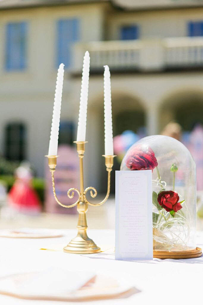Table setting with a gold three-candle candelabra, a menu, and a glass cloche with red faux flowers.