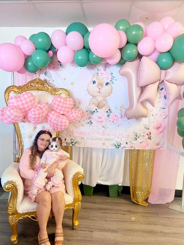 Mother holding baby girl seated in gold ornate throne chair before Some Bunny is Turning One birthday backdrop