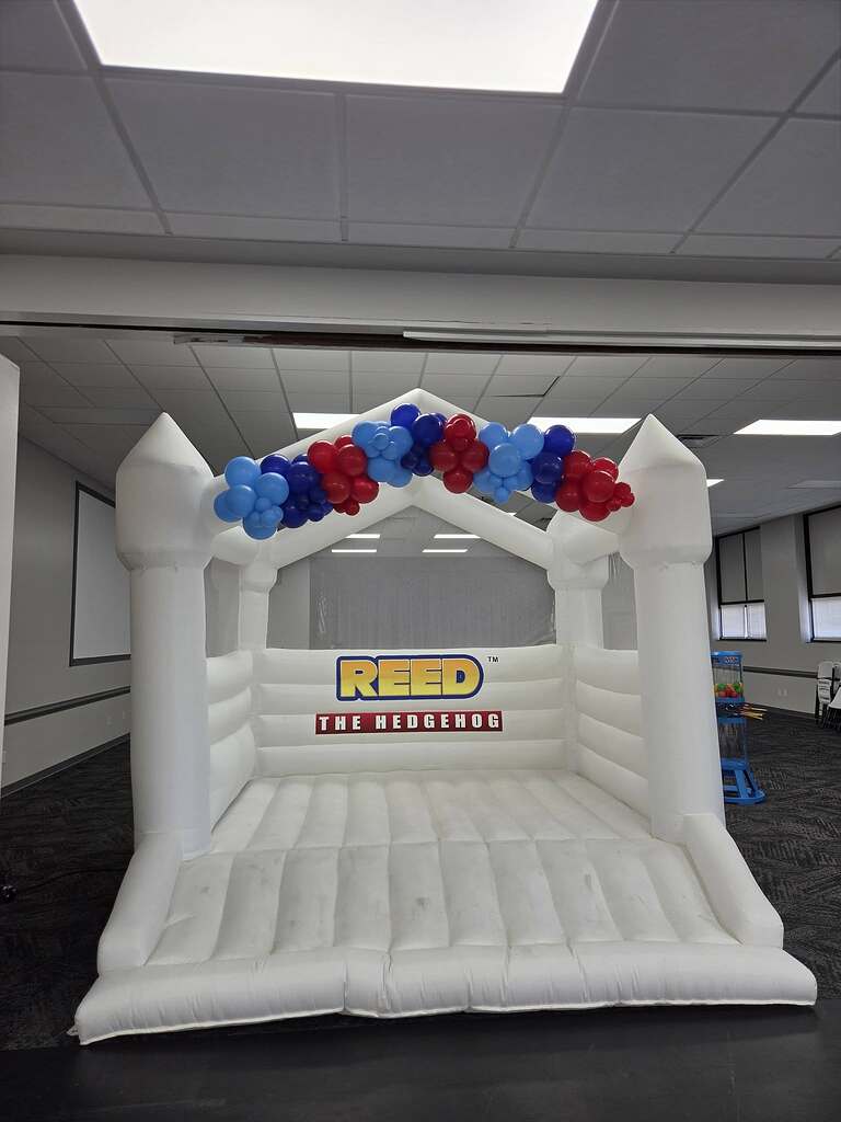 White inflatable bounce house branded with Reed The Hedgehog and decorated with red and blue balloons