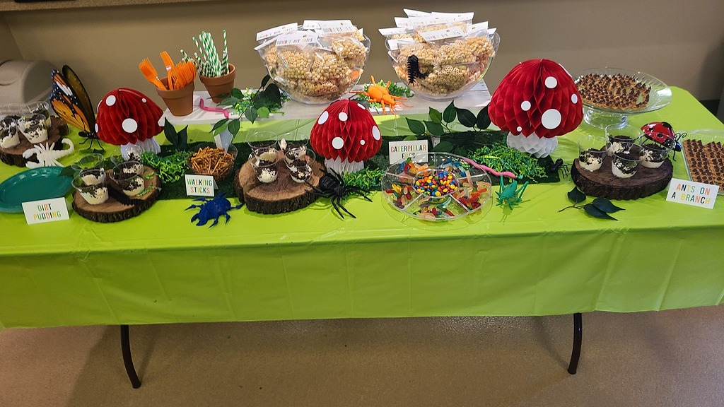Table decorated with an insect-themed party spread featuring toadstools, dirt pudding, and bug candy.