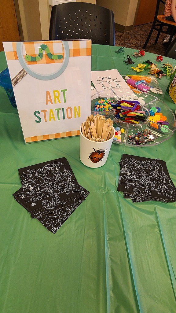 Table with an Art Station sign, craft supplies including popsicle sticks, beads, and black insect-themed coloring cards.