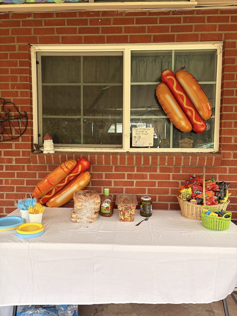 A party table decorated for a hot dog theme with balloon hot dogs, snacks, and The Pigeon Finds a Hot Dog sign.