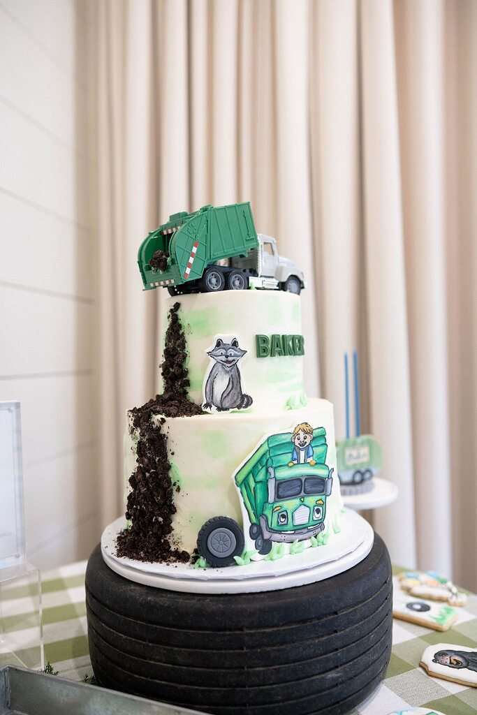 Two-tiered garbage truck themed birthday cake with a raccoon decoration and edible cookie crumbs