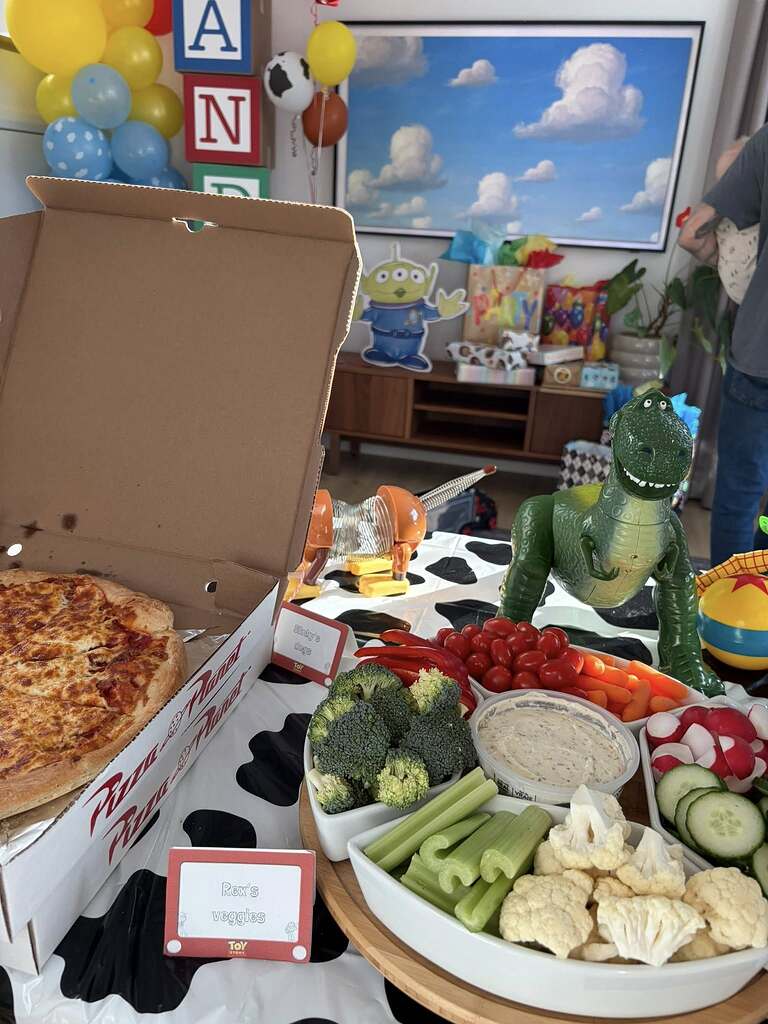 Toy Story themed party table with Pizza Planet pizza box, assorted fresh raw vegetable platter, and Rex and Slinky Dog figures.