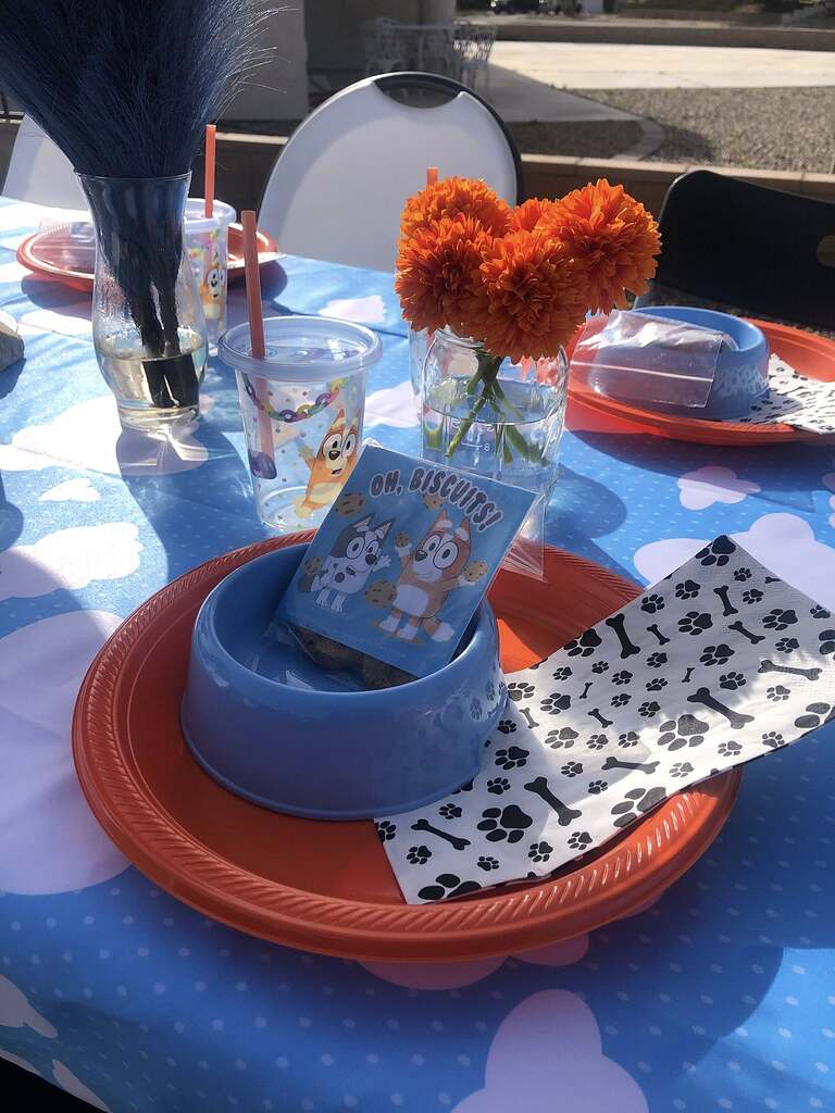 Bluey-themed party place setting with a light blue bowl, paw print napkin, and biscuit bag on an orange plate.