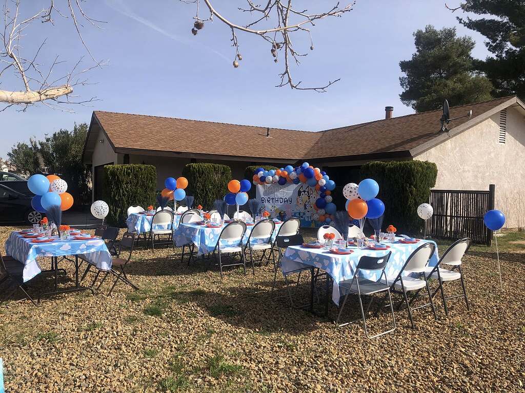 Outdoor birthday party setup with blue cloud-patterned tablecloths and blue and orange balloon arches.