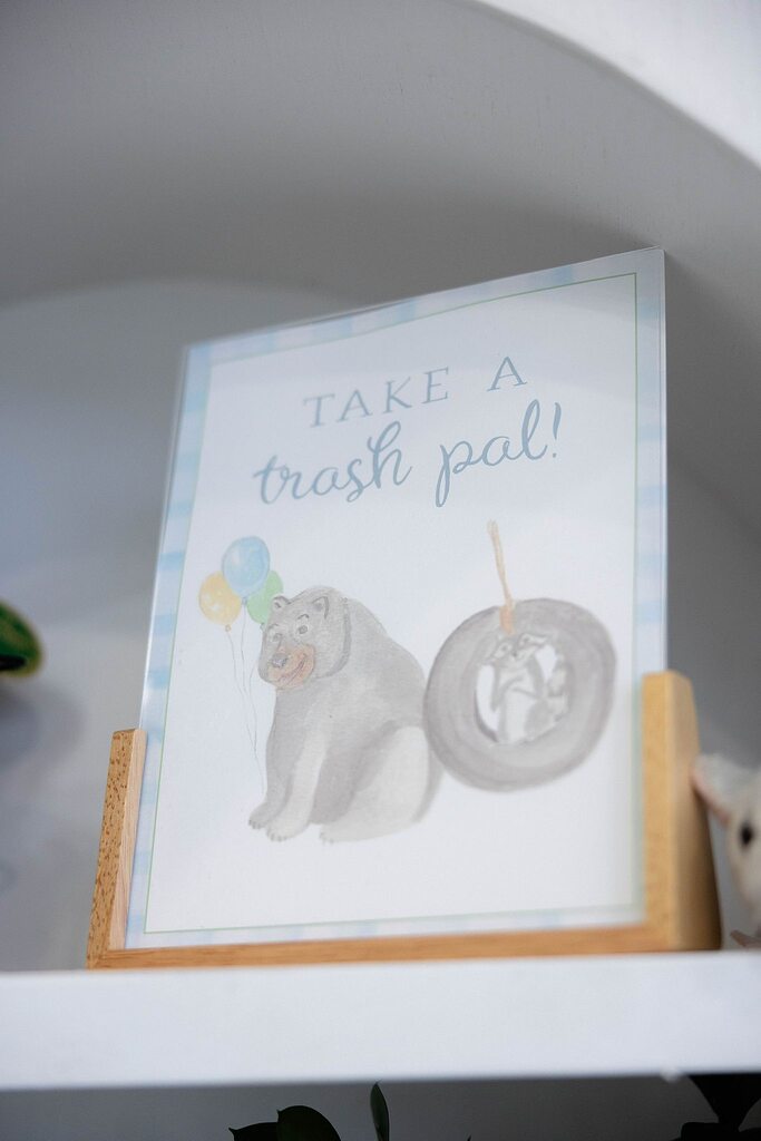 Table sign featuring a watercolor illustration of a bear with balloons and a raccoon in a tire labeled Take a trash pal