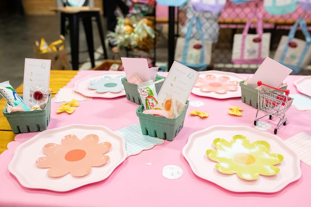 Table setting with flower-shaped plates, fruit-themed party favors in green baskets, and a mini shopping cart.