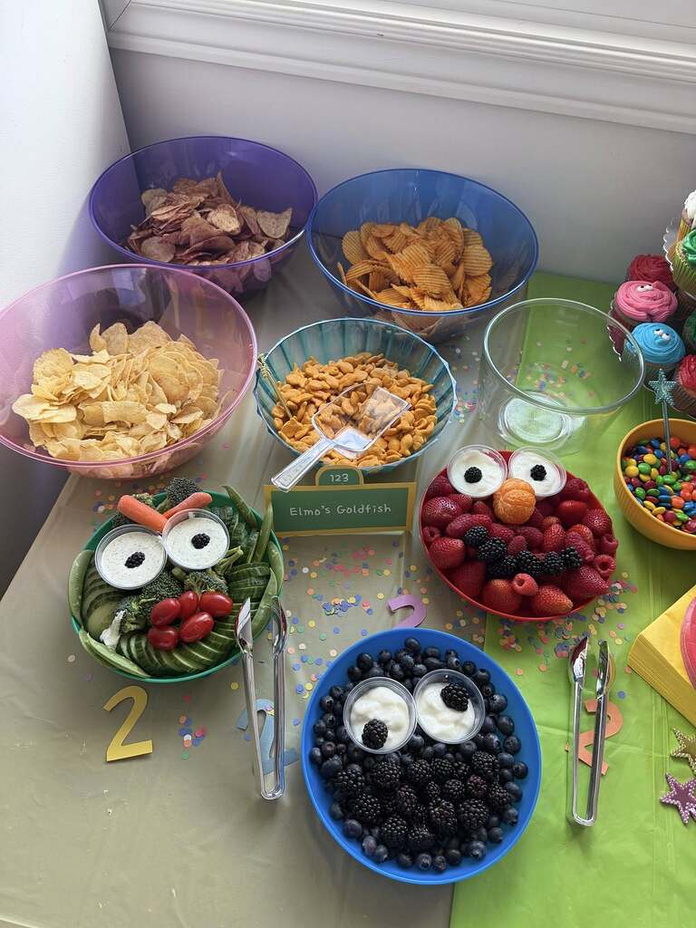 Sesame Street-themed party food table with bowls of chips and snack trays shaped like Cookie Monster and Elmo.