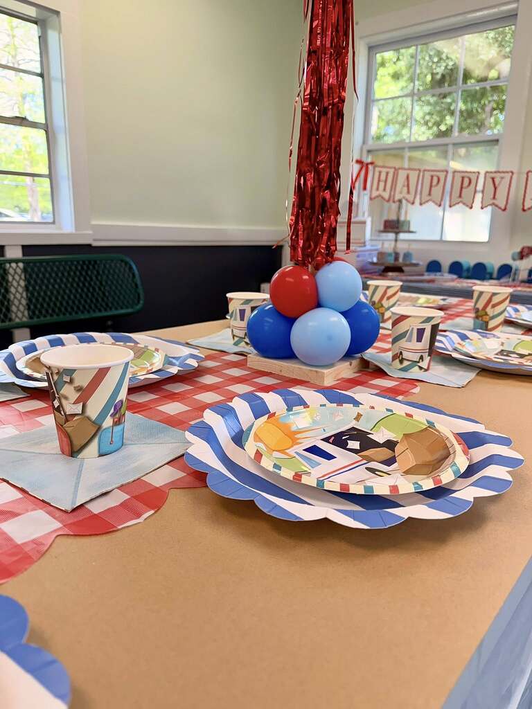 Mail carrier themed party table setting with patterned paper plates, cups, and red, white, and blue balloons.