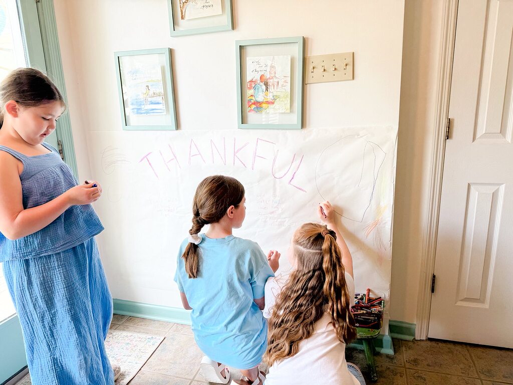 Three young girls coloring and writing on a large sheet of paper taped to the wall labeled Thankful.