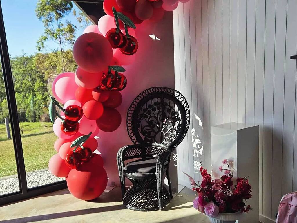 Black peacock chair next to a red and pink balloon garland with cherry-shaped foil balloons.