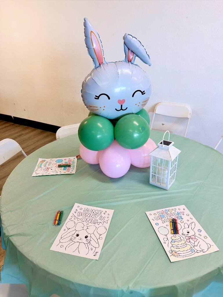Table set for a first birthday party with a bunny balloon centerpiece, coloring sheets, and crayons.