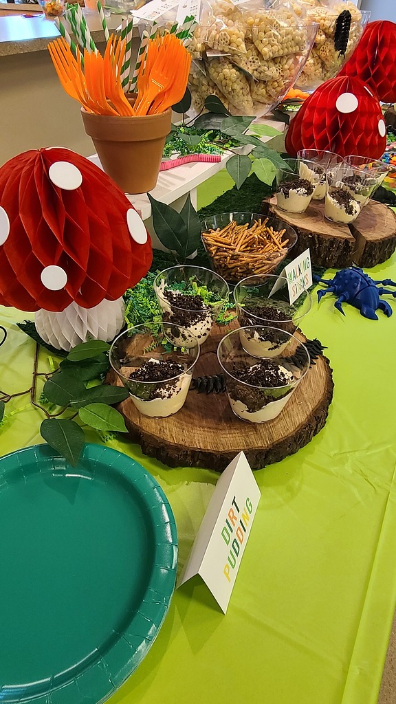 Forest-themed party table featuring dirt pudding cups, pretzel 'walking sticks', and mushroom decorations.