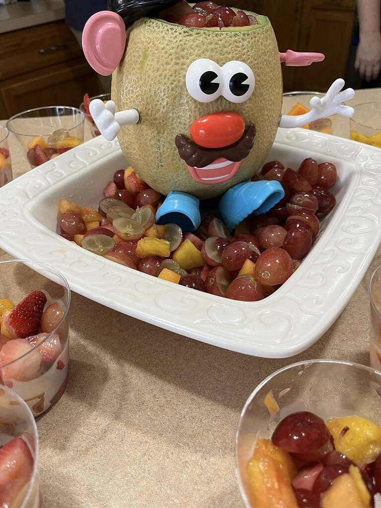 Cantaloupe carved into a Mr. Potato Head shape sitting in a bowl of fresh fruit salad.