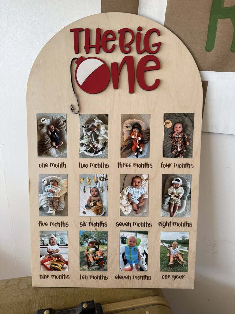 Wooden arch sign displaying The Big One fishing theme milestone board with monthly baby photos from one month to one year.