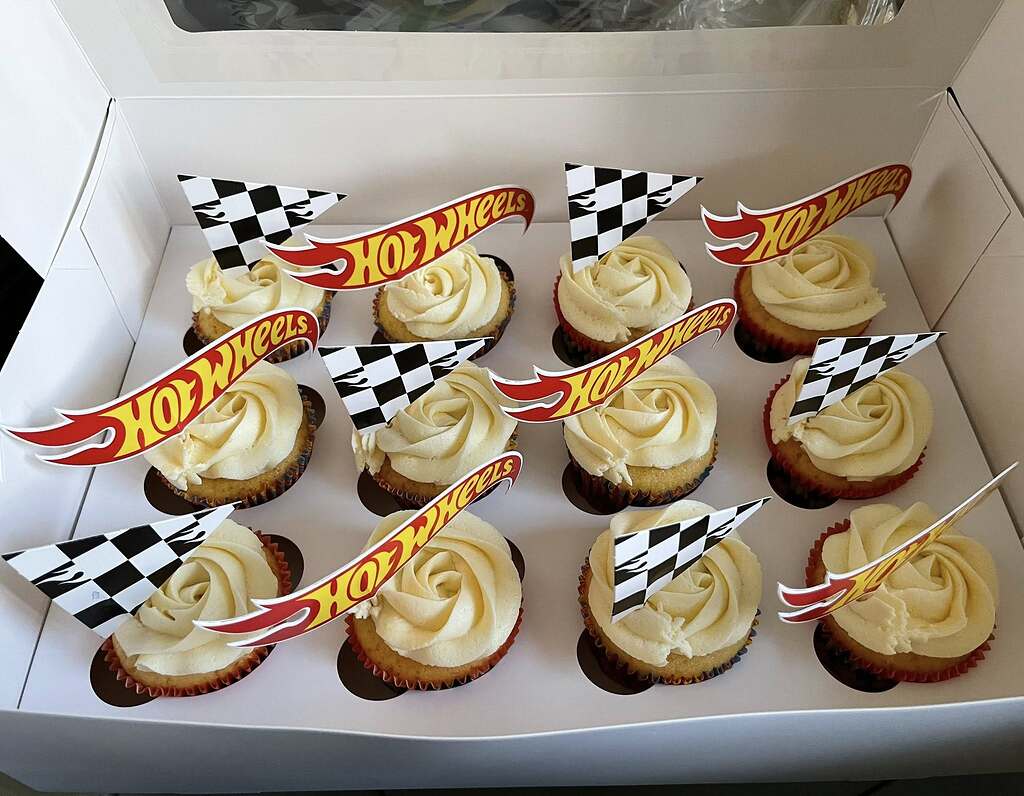 Dozen vanilla cupcakes with buttercream frosting, decorated with Hot Wheels checkered flags and logo toppers.