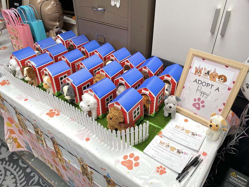Adopt a Puppy party favor display featuring plush puppies in individual red cardboard dog houses with white picket fencing and adoption certificates.
