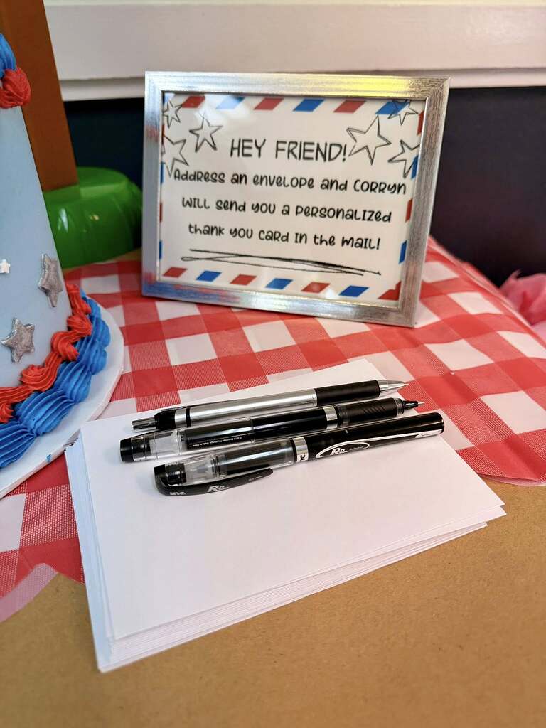 Table sign inviting guests to address an envelope for a personalized thank you card, with pens and stationery.