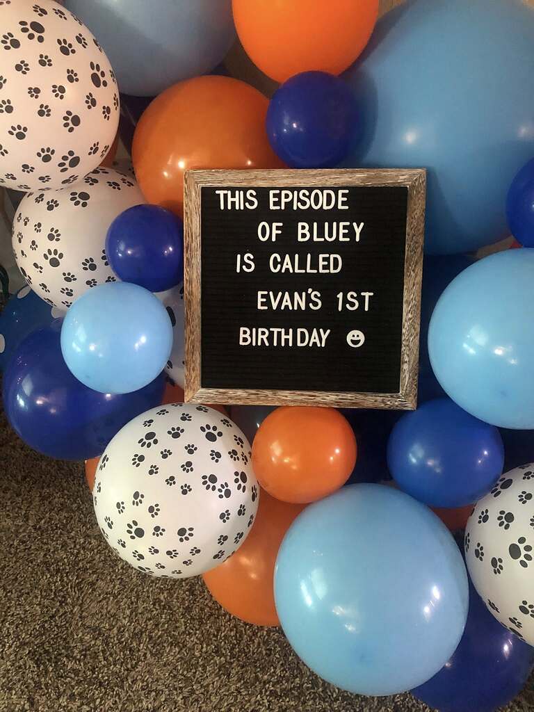 Bluey-themed first birthday party sign surrounded by orange, blue, and paw print balloons.