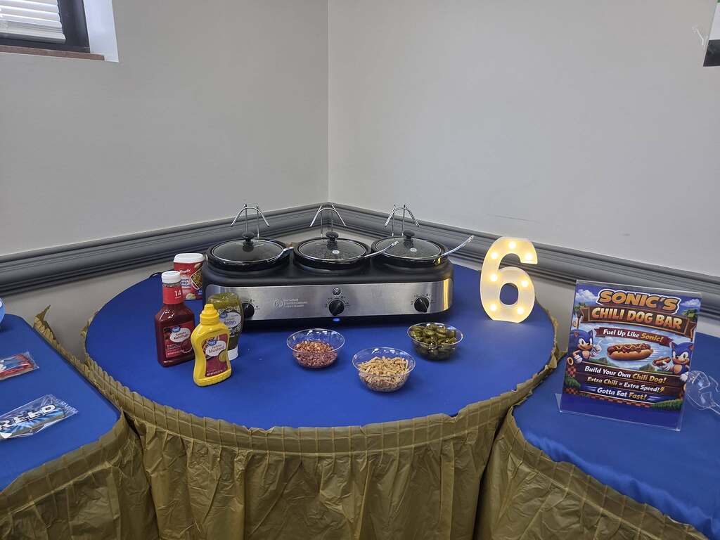 Sonic the Hedgehog themed chili dog bar setup with warming pots, condiments, and toppings on a blue tablecloth.