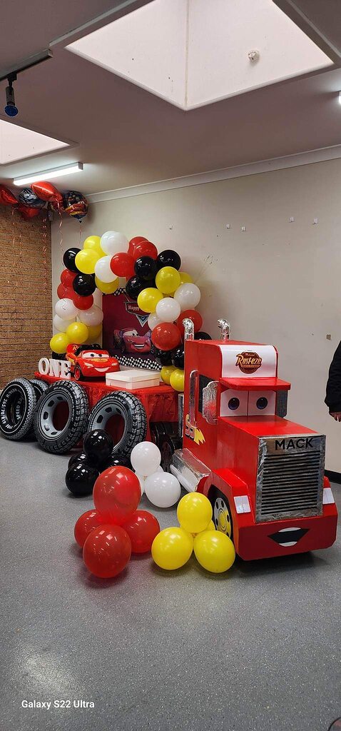 Cars themed first birthday party decoration featuring a large Mack truck prop, tire accents, and colorful balloons.