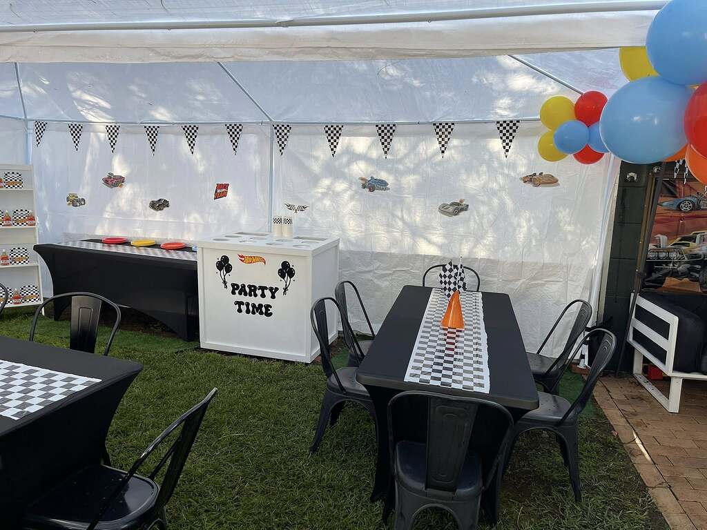 Hot Wheels themed party setup with checkered table runners, race car wall decorations, and colorful balloon clusters.