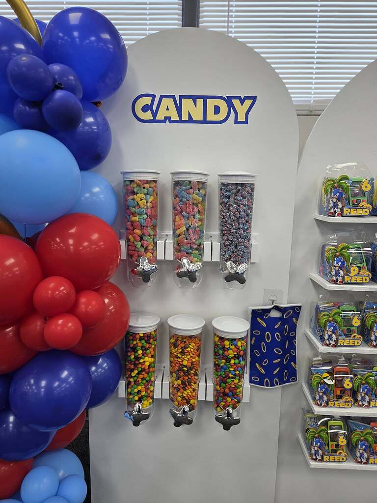 Candy dispenser station with rows of colorful gummies and chocolates decorated with blue and red balloons.