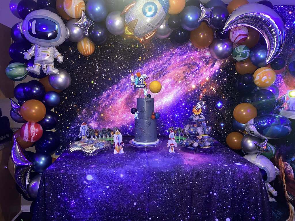 Space themed birthday dessert table with galaxy backdrop and balloon arch featuring planets and astronaut decor.