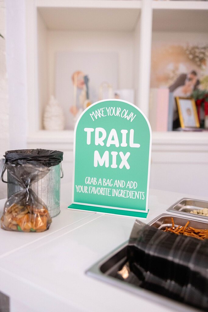 Table sign for a DIY trail mix bar with ingredients in trays and a container of bags.