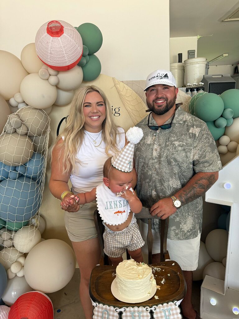 Parents posing with their one-year-old baby at a first birthday party with a fishing-themed cake and balloons.