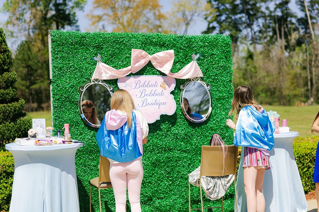 Two people wearing blue capes standing in front of a Bibbidi Bobbidi Boutique backdrop decorated with mirrors.