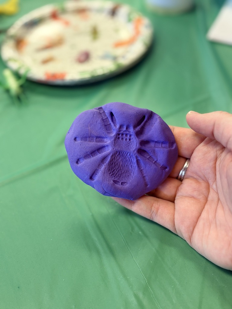 Hand holding a piece of purple modeling clay featuring a stamped spider impression on a green surface.