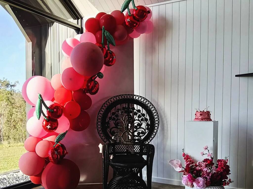 Birthday party setup with cherry balloon garland, black peacock chair, and small pink cake on a pedestal.