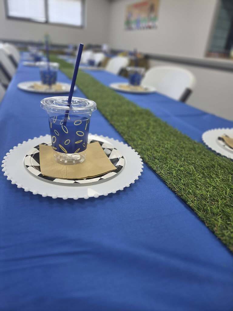Table setting with blue tablecloth, grass runner, charger plate, and patterned cup with straw.