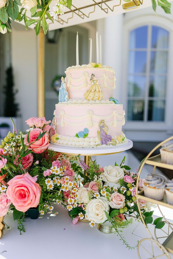 Two-tier pink princess birthday cake with Disney princess toppers surrounded by floral arrangements.