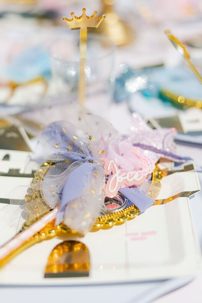 Table setting with gold crown decor elements, star-patterned ribbon, and a customized name tag on a party plate.