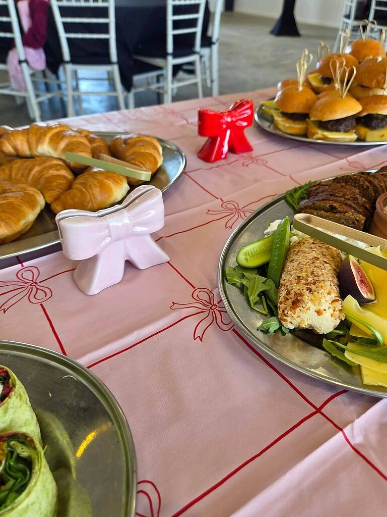 Table spread showcasing croissants, sliders, and a charcuterie platter on a pink bow-patterned tablecloth.