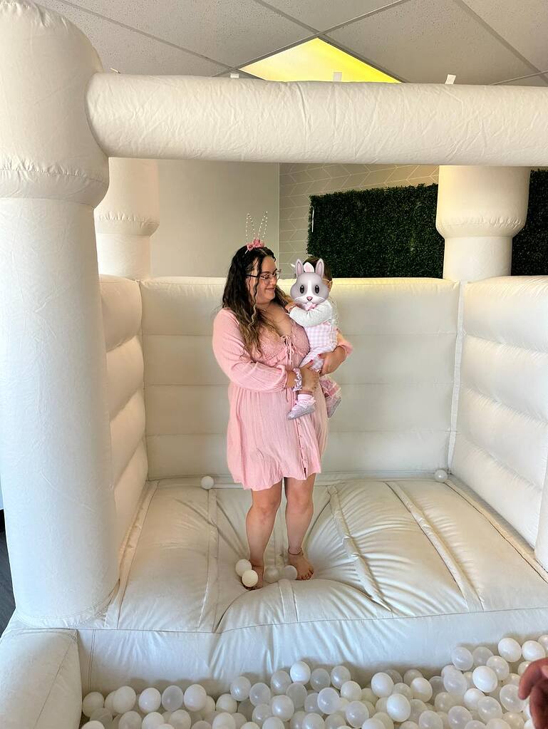Woman in a pink dress holding a toddler inside a white bounce house with ball pit balls.