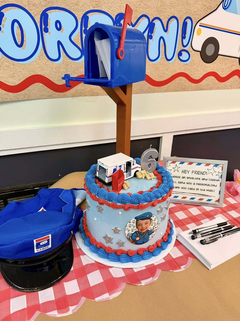 Mail carrier themed birthday cake with a number 6 candle, toy mail truck, and photo topper on a table with a postal hat.