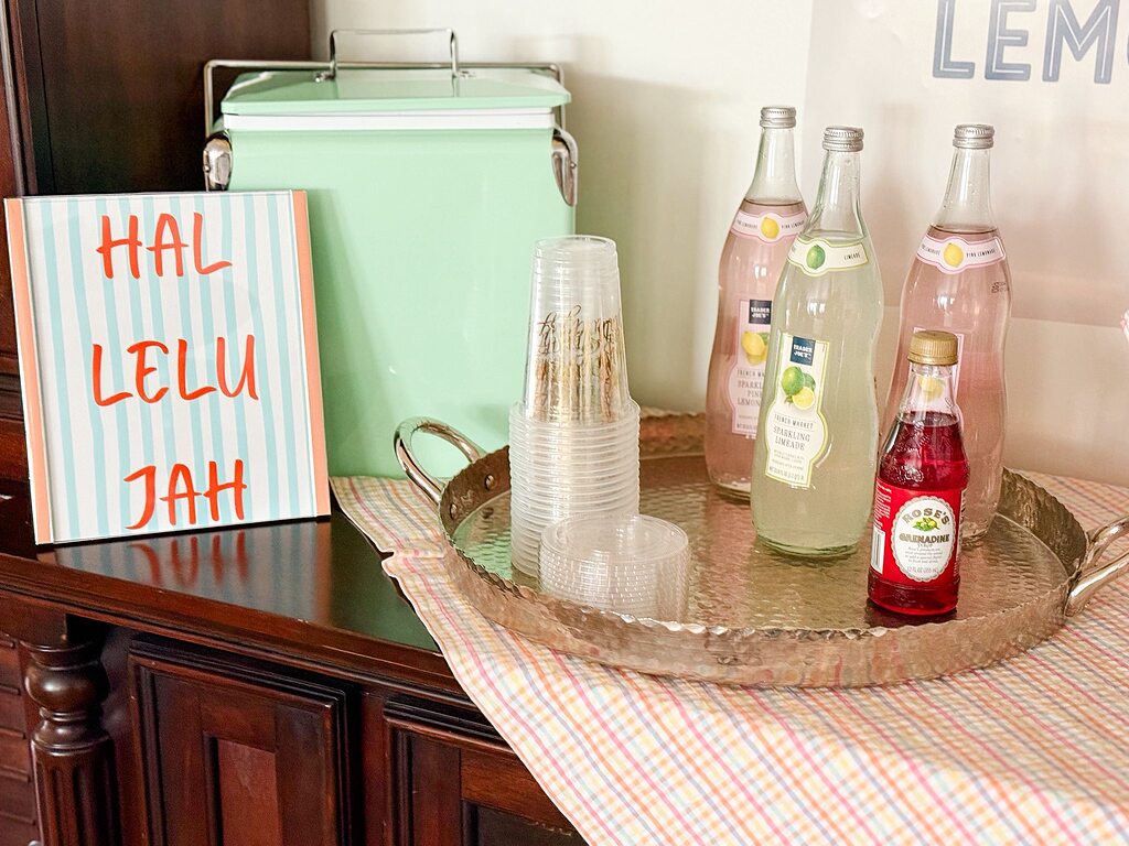 Drinks station featuring bottles of sparkling limeade and grenadine, stacked plastic cups, and a mint green cooler.