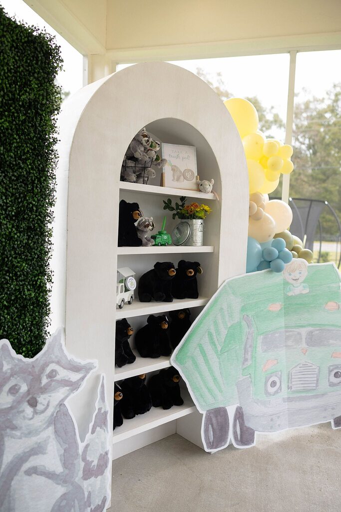 White arched display shelf filled with plush black bears and raccoon toys with a Take a Trash Pal sign