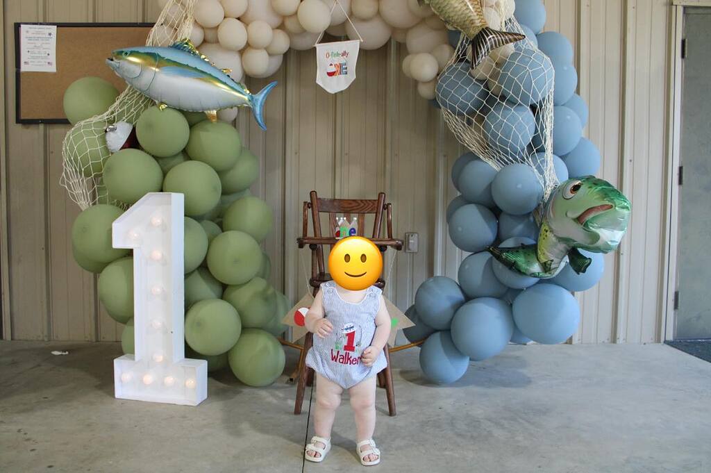 First birthday backdrop with balloon arch, fishing nets, fish balloons, and a number one marquee light.