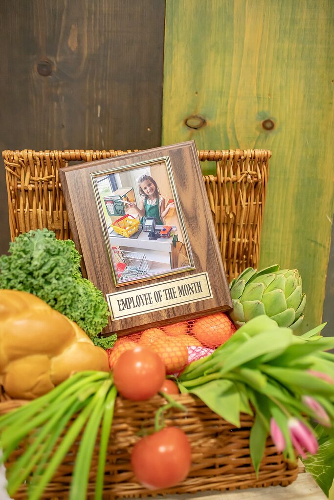 Wicker basket filled with fresh produce and an Employee of the Month award frame showing a girl playing cashier.