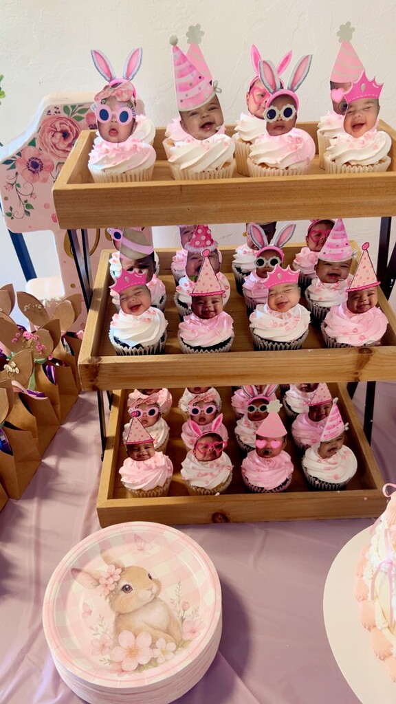 Three-tier wooden stand filled with cupcakes decorated with pink icing and custom baby photo toppers.