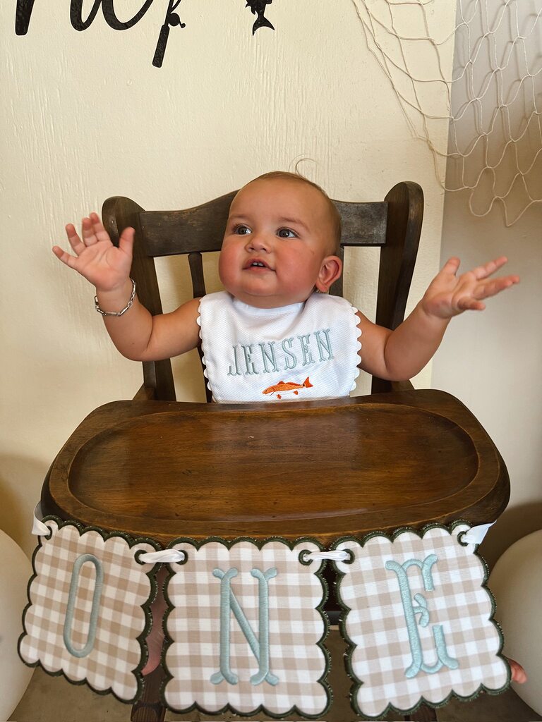 Baby sitting in a wooden high chair wearing a personalized white bib with the name JENSEN and an embroidered fish